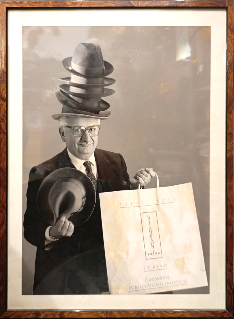 Bruno Gambarotta Fotografia di Bruno Gambarotta con un cappello in mano, una busta della Cappelleria Viarani e più cappelli impilati sulla testa.